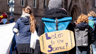 Person am Demo mit Schild am Rücken. Darauf steht 'Listen to the Science!'